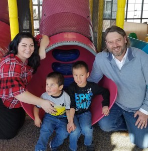 A family of four smiling at the camera. Two little boys sitting at the bottom of a slide with mom and dad on either side.