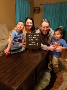 A family of four smiling to the camera. A sign in front of them says, "We've waited our whole lives for this moment."
