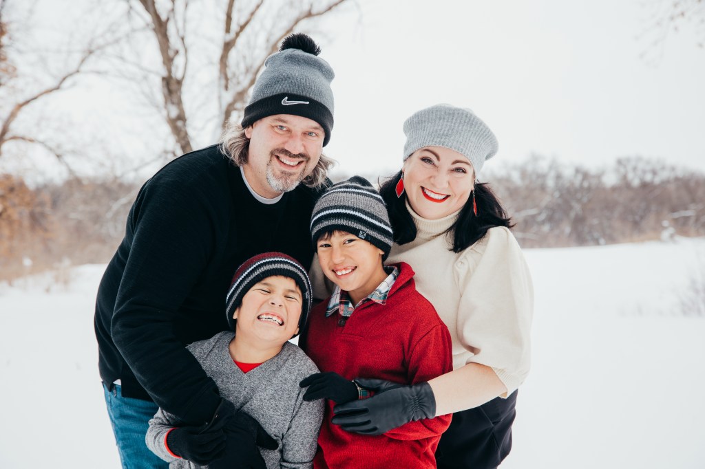 A family of four with a dad, mom, and two boys. Standing and smiling in a snowy field wearing hats, gloves, and sweaters.
