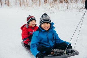 Two boys on a sled being pulled by a sting. The boy in the front is smiling at the camera.