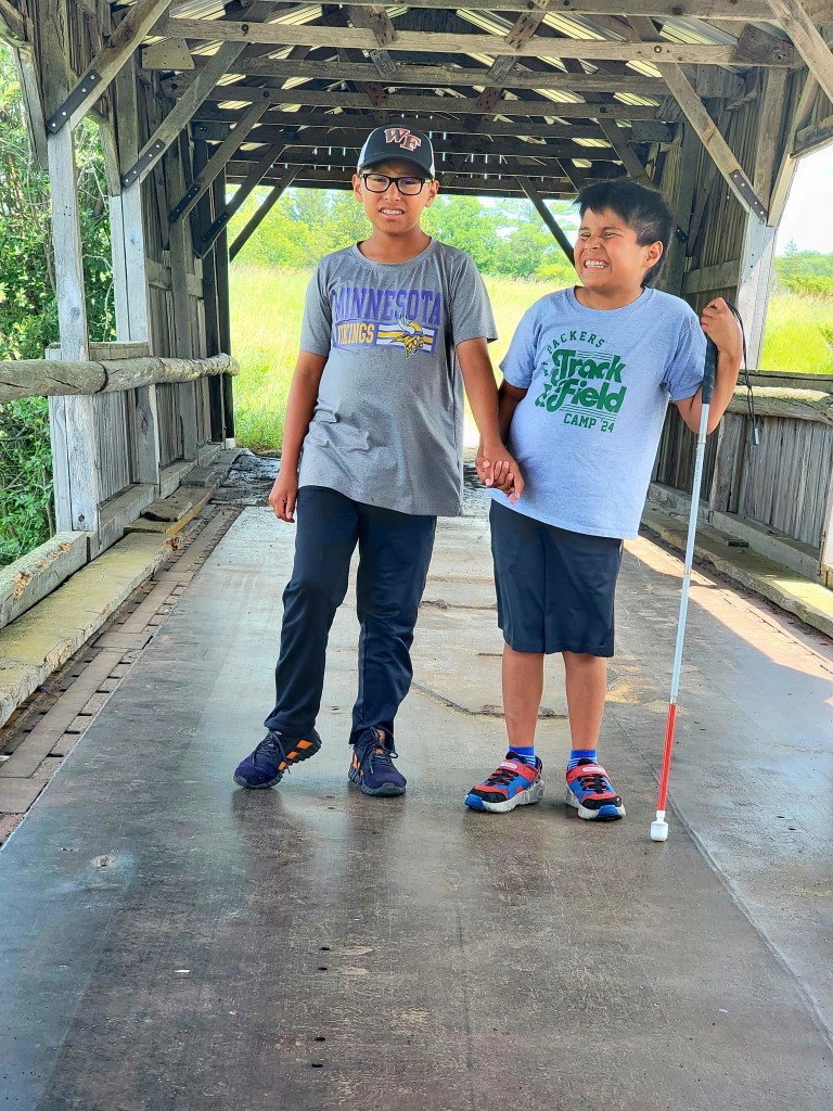 Two boys smiling on a covered bridge, one boy hold a white cane.