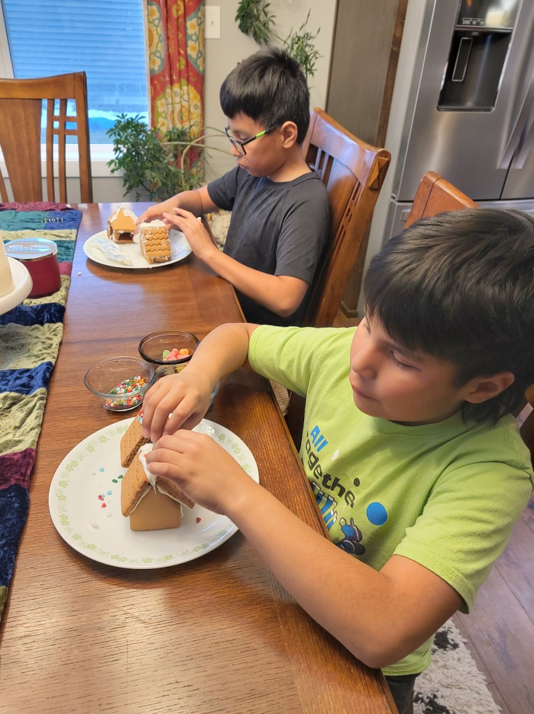 Two boys sitting at a table adding candies to their gingerbread houses.