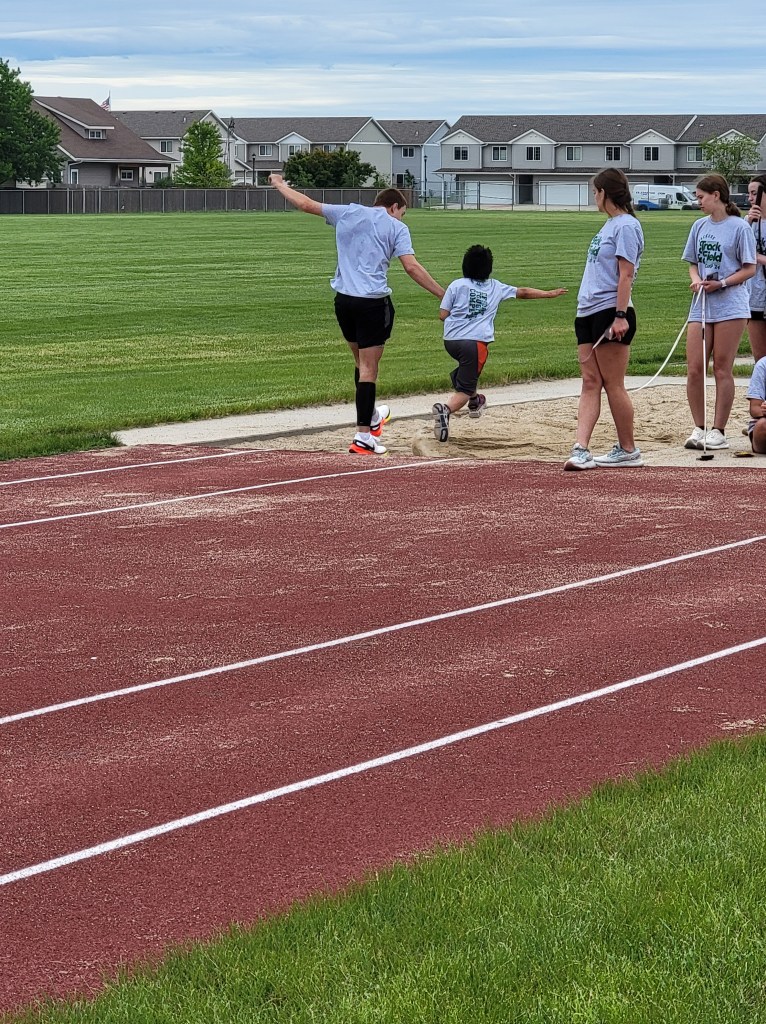 A young boy doing a long jump into a sand pit while an older boy is holding his hand guiding him. A older girl watches.