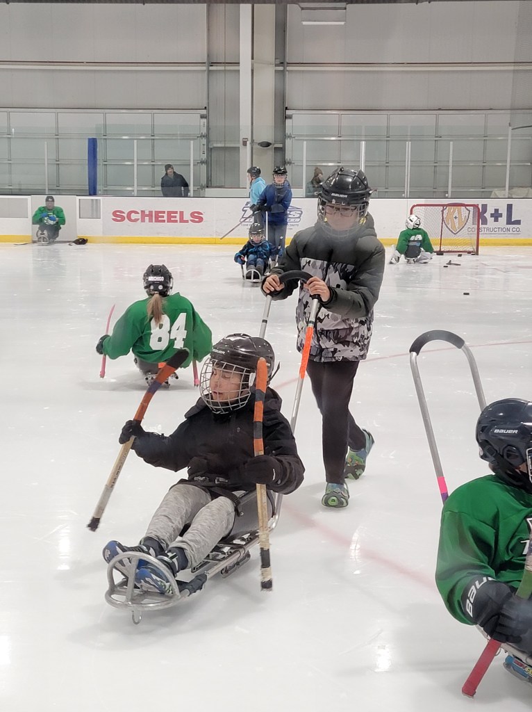 One boy lpushing another boy on a sled. They at a hockey rink with other sled hockey players in the background.