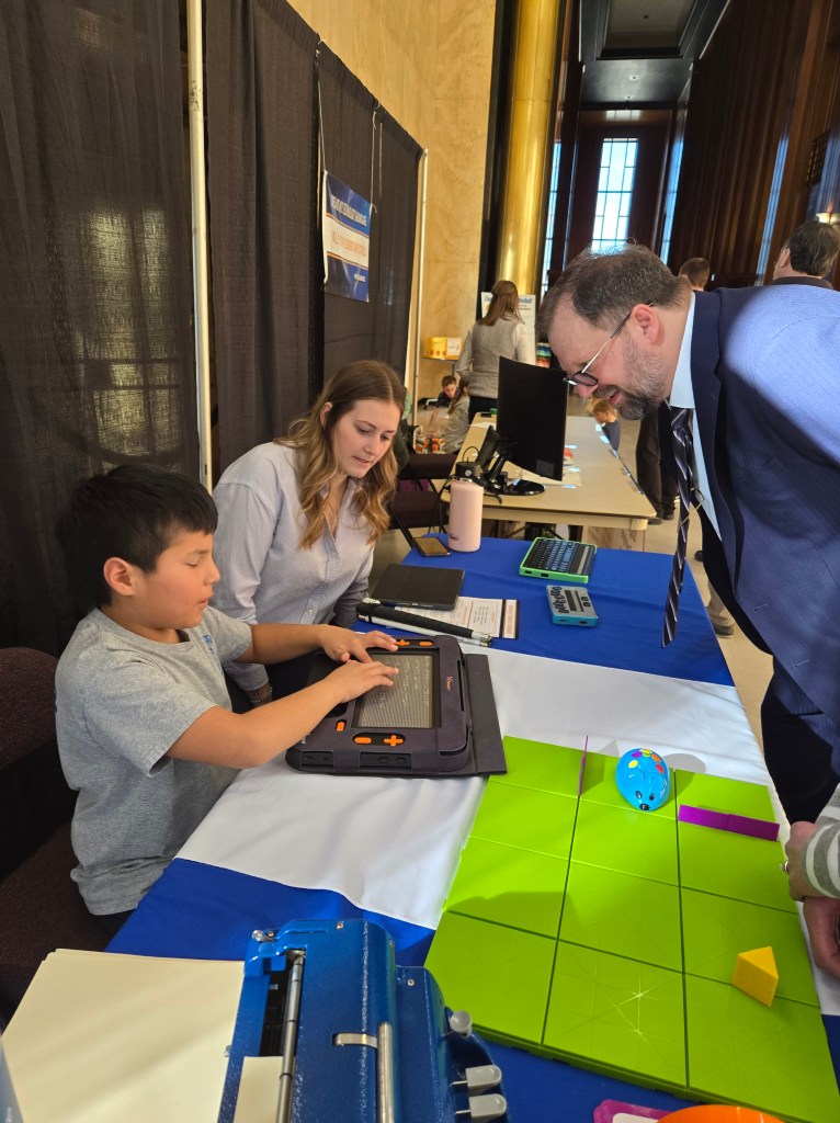 A young boy sitting behind a table demonstrating how the Monarch works. Using his fingers to read the braille dots, while others look on. 