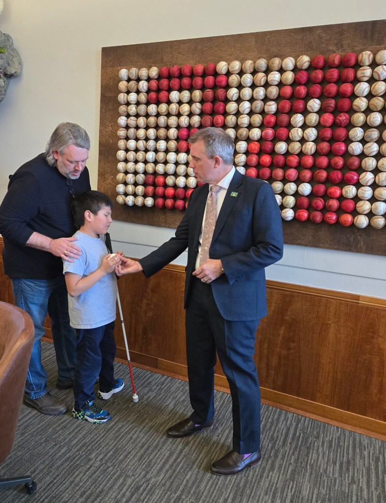 A young boy holding a white cane receiving a baseball from the governor. 