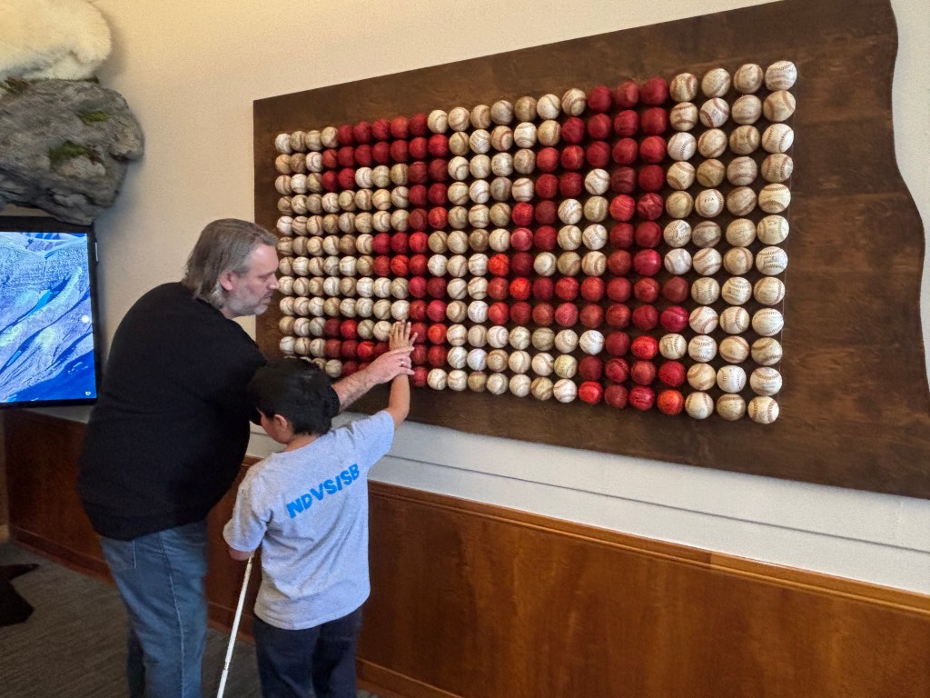 A young boy and his father standing in front of a giant baseball art work looking at the balls with his hands. 