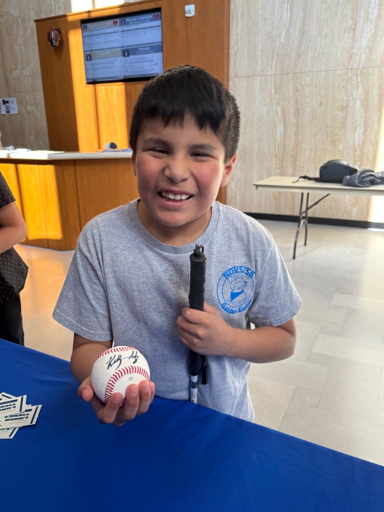 A young boy holding a white cane smiling big showing his signed baseball in the other hand. 