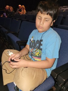 A little boy holding the receiver with an earpiece in his ear, sitting in a chair in a theater. 