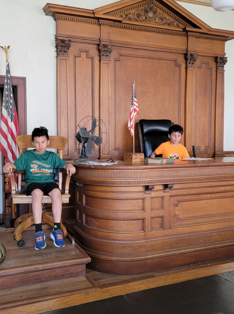Old courthouse with a large desk. One boy is sitting in the judges chair while another boy is in the witness chair next to the desk. The American flag is beside the desk. 