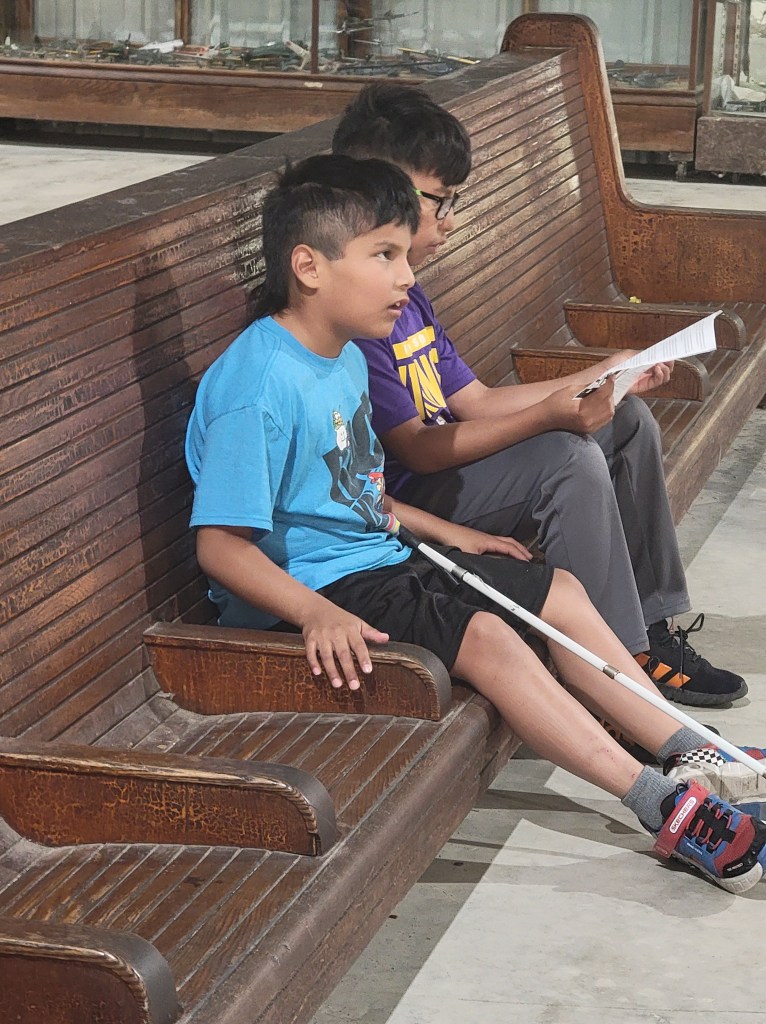 Two young boys sitting on a long wooden old time bench waiting for their plane to arrive. One boy has a white cane in his lap while the other boy is looking at a paper. 