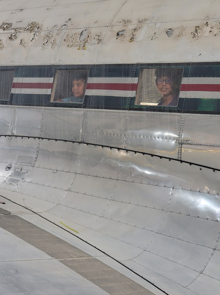 Outside of an old plane focusing on two windows. On the other side of the windows looking out are two boys, smiling at the camera. 