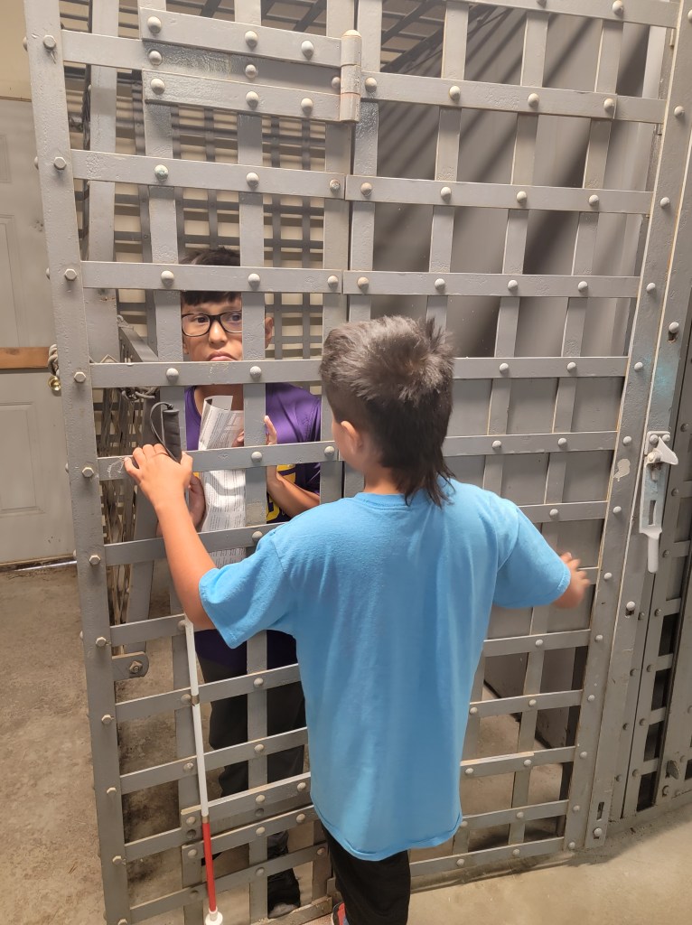 Little boy standing outside a old time jail cell, holding a cane in his left hand locking in an older boy. 