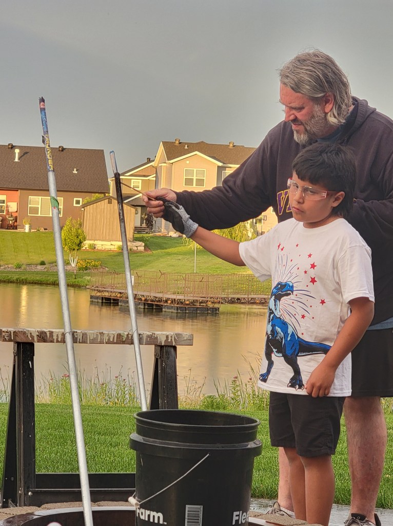 Young boy wearing safety glasses and a red, white, and blue dinosaur shirt. A man holding the hand of the boy with a punk. lighting a bottle rocket. 