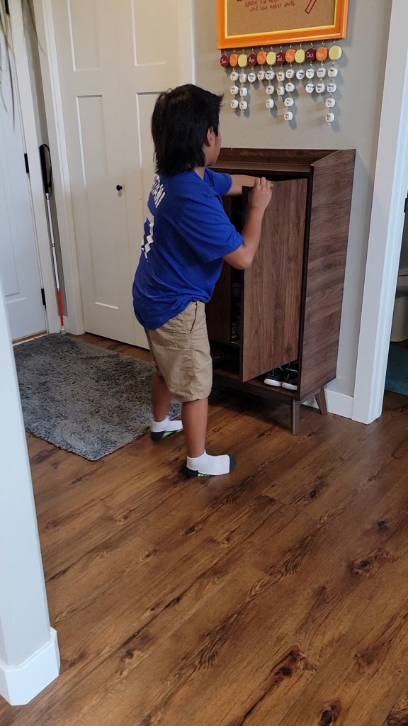 A young boy opening a wooden cabinet and reaching in to get his shoes. He is wearing a Dodger blue Ohtani 17 t-shirt and shorts. 