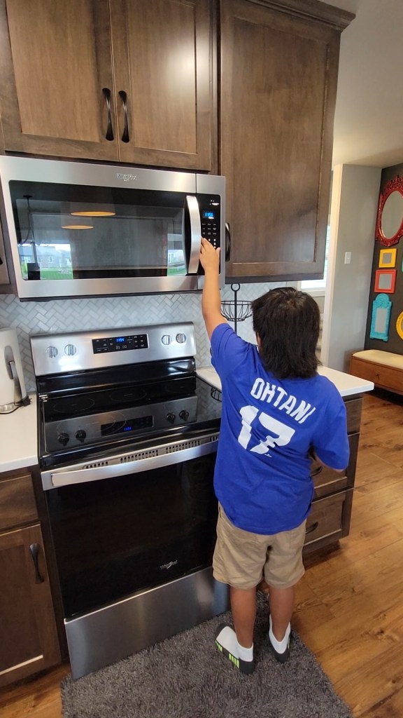 A young boy wearing a Dodger blue Ohtani 17 t-shirt is toughing clear bump dots on the microwave. He is standing on a small rug in front of the microwave.