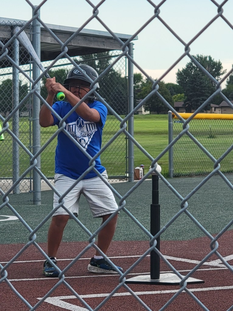 Boy wearing blue shirt. grey shorts, and a batting helmet in mid swing.