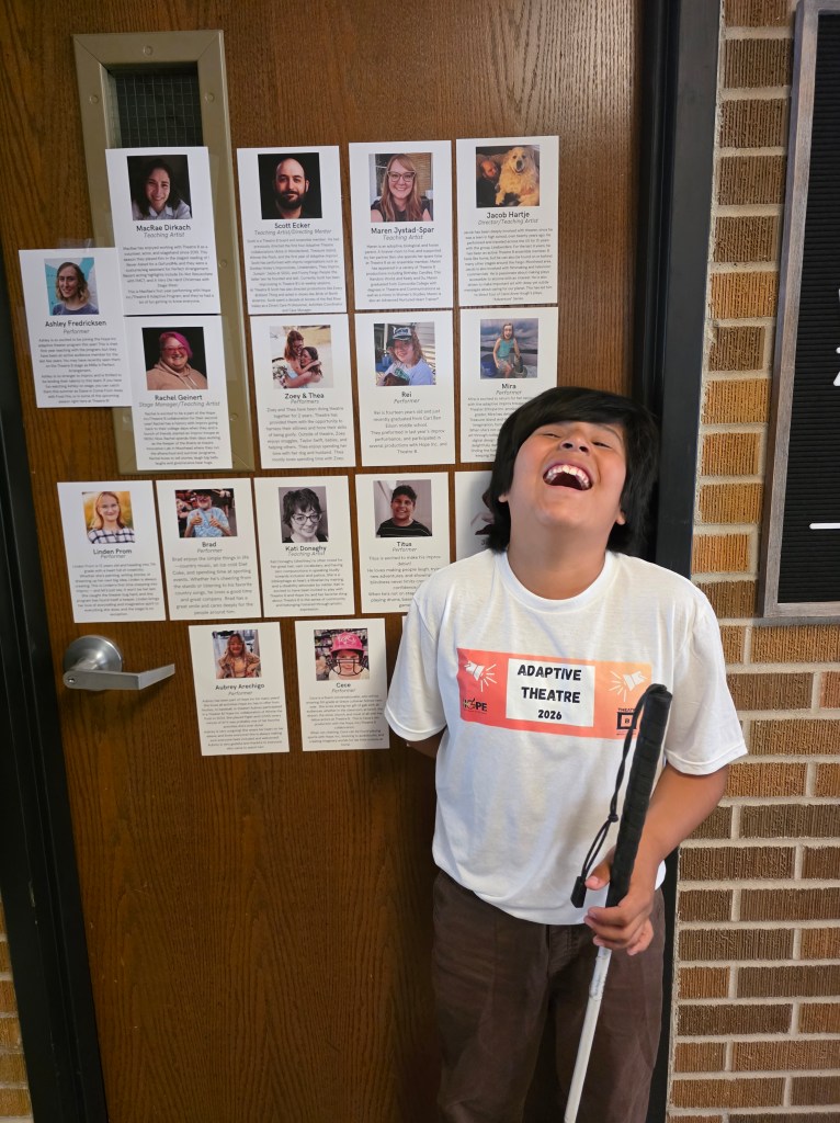 A little boy holding a white cane smiling big. He is wearing an Adaptive Theatre 2026 shirt and he is standing in front of a door showcasing all the performers, including his own picture and write-up.