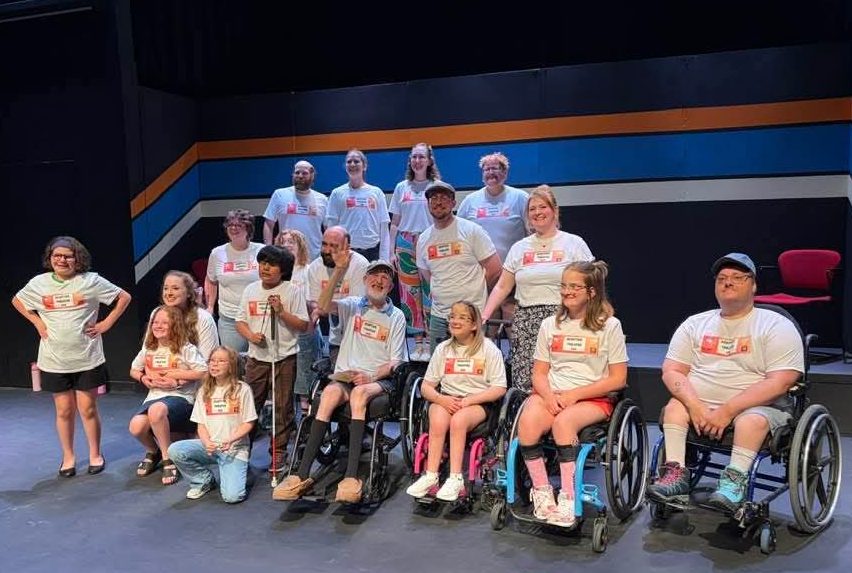 A group of 18 performers all wearing Adaptive Theatre 2026 shirts. They are on and in front of the stage. Some are standing, some are kneeling, and some are sitting in wheelchairs. All performers are smiling. 