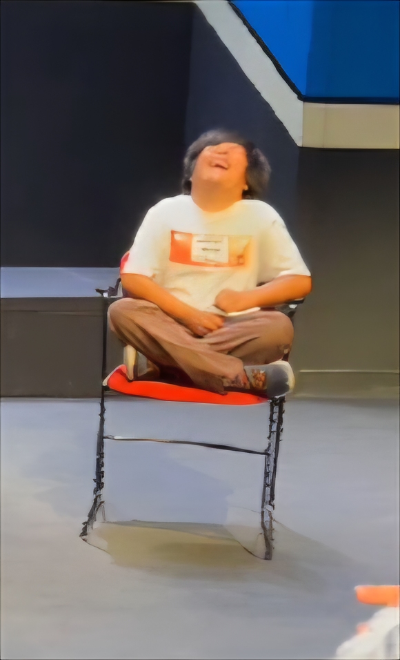 A little boy sitting cross legged on a chair, on a stage. His head is flung back in the throws of a huge laugh. 