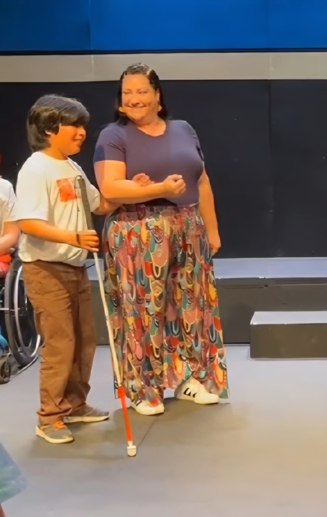 A little boy holding a white cane being guided on a stage by a woman. Both are smiling. 
