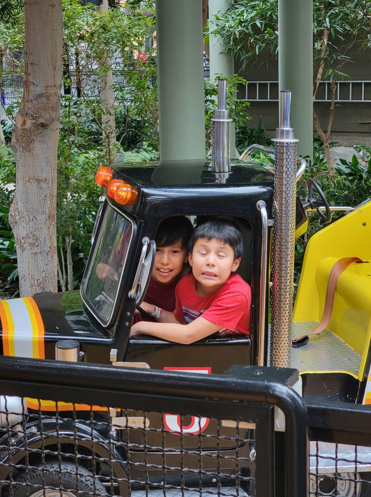 Two boys looking at the camera sitting inside a small semi truck amusement park ride. 